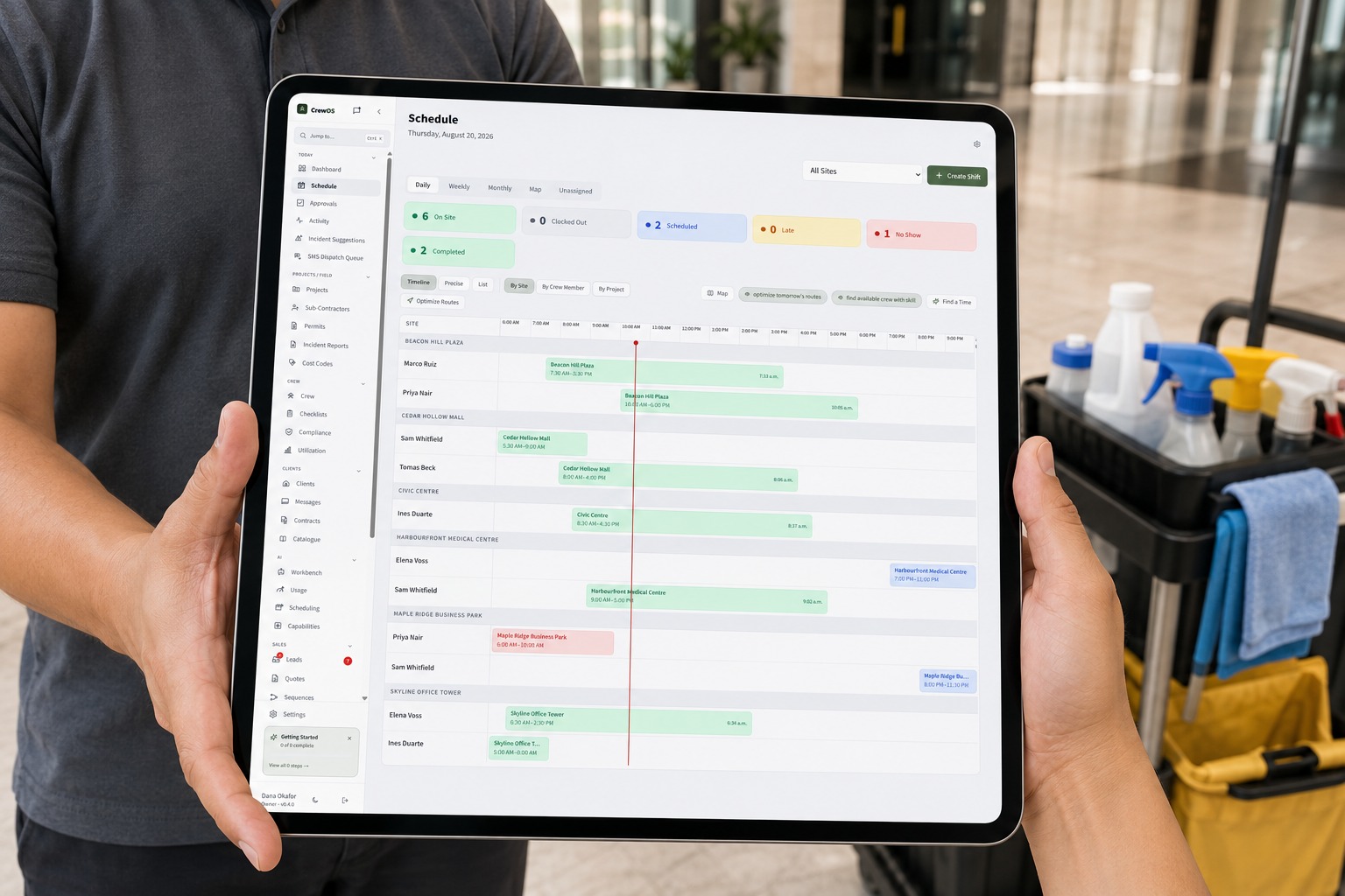 A commercial cleaner with a cleaning cart in an office lobby holding a tablet that shows the CrewOS schedule with live status across every property