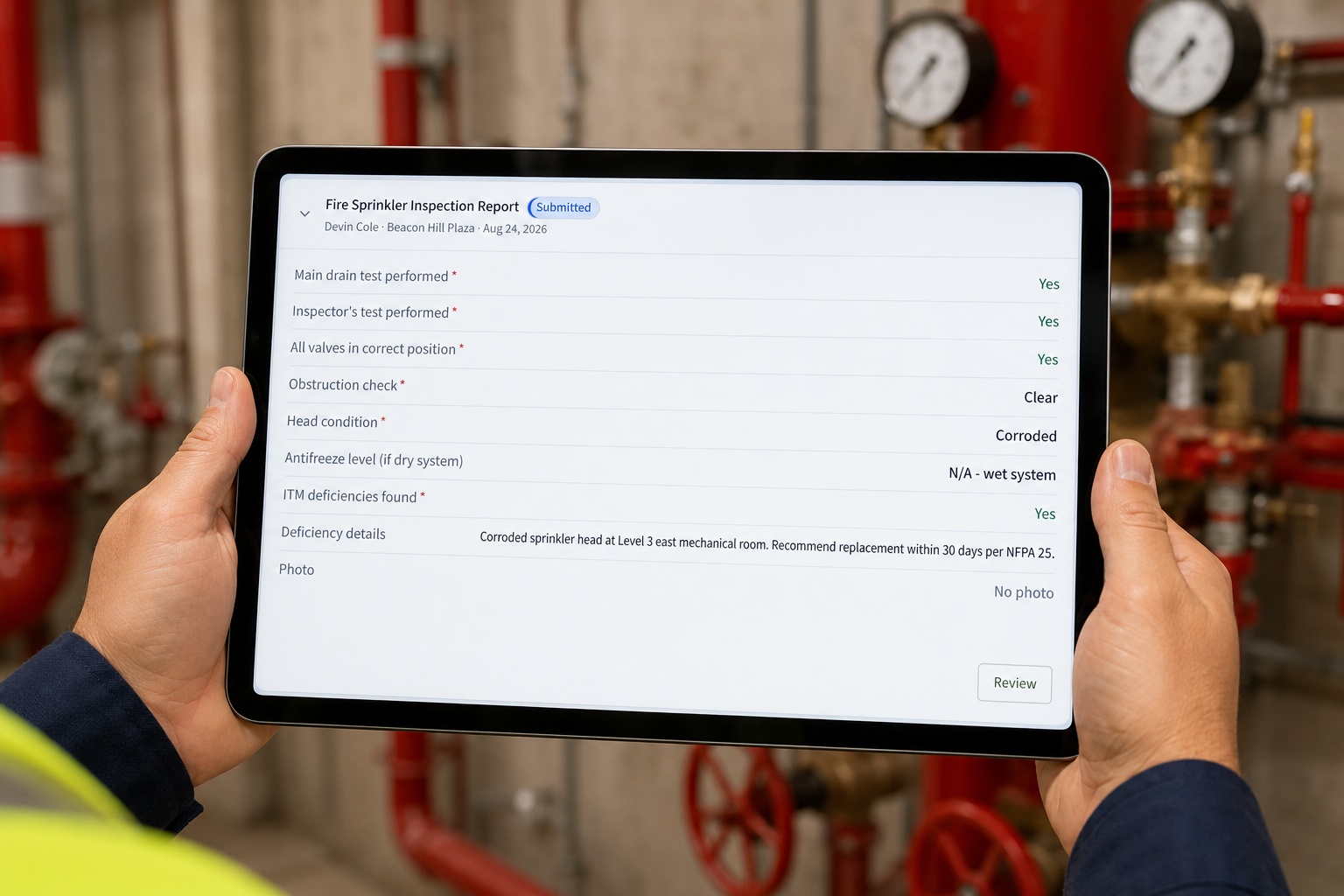 A fire protection inspector in a hard hat holding a tablet that shows a CrewOS fire sprinkler inspection report, standing at a red sprinkler riser in a building mechanical room