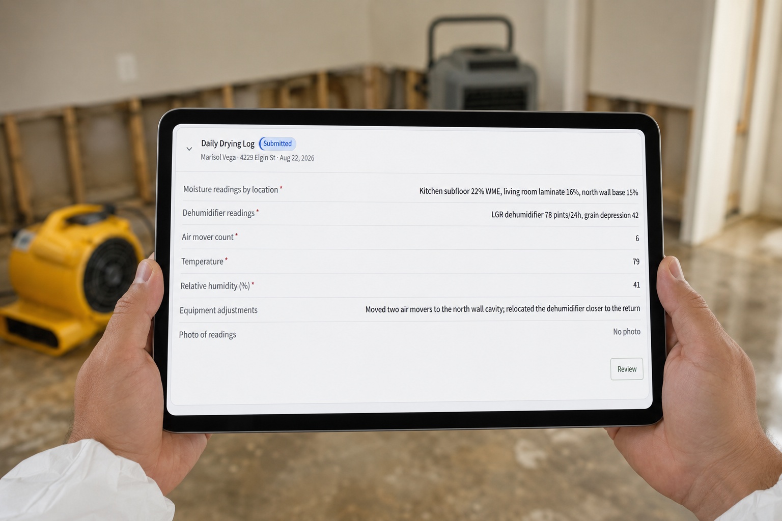A restoration technician holding a tablet that shows a CrewOS daily drying log, standing in a water-damaged room with air movers and a dehumidifier running