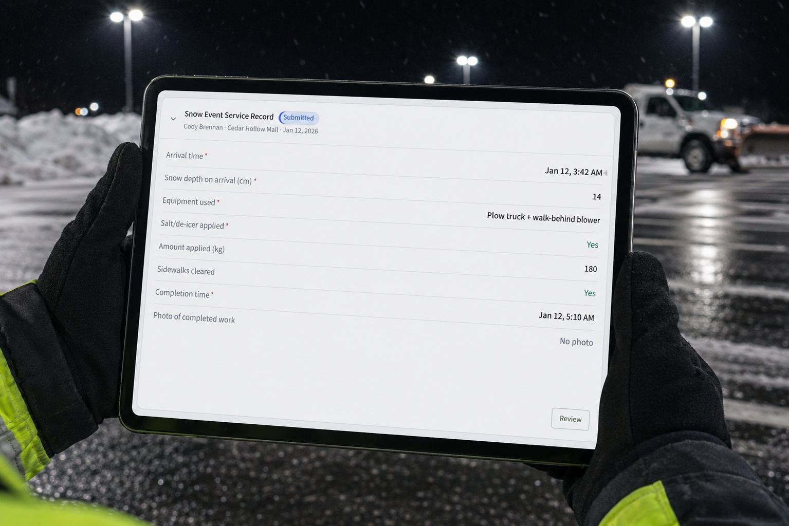 A snow removal operator holding a tablet that shows a CrewOS snow-event service record, standing in a cleared commercial parking lot at night with a plow truck and snow piles behind